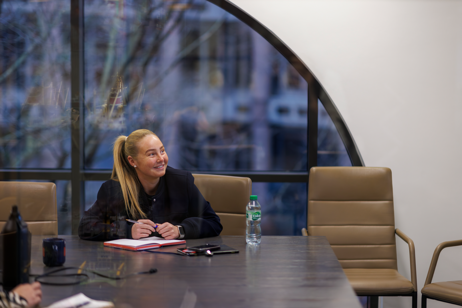 Smiling woman in a black blazer sitting at a conference table with a notepad.