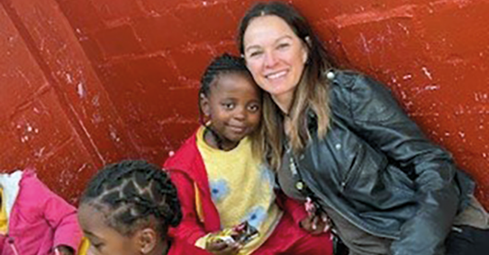 Woman in leather jacket sitting next to a young girl in a colourful outfit against a red wall.