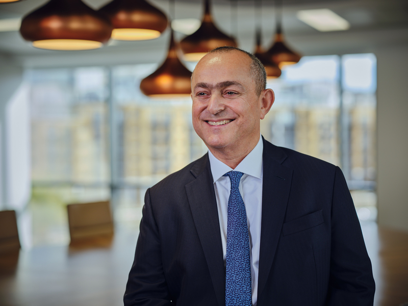 Smiling man in a dark suit standing indoors with a cityscape view through large windows.