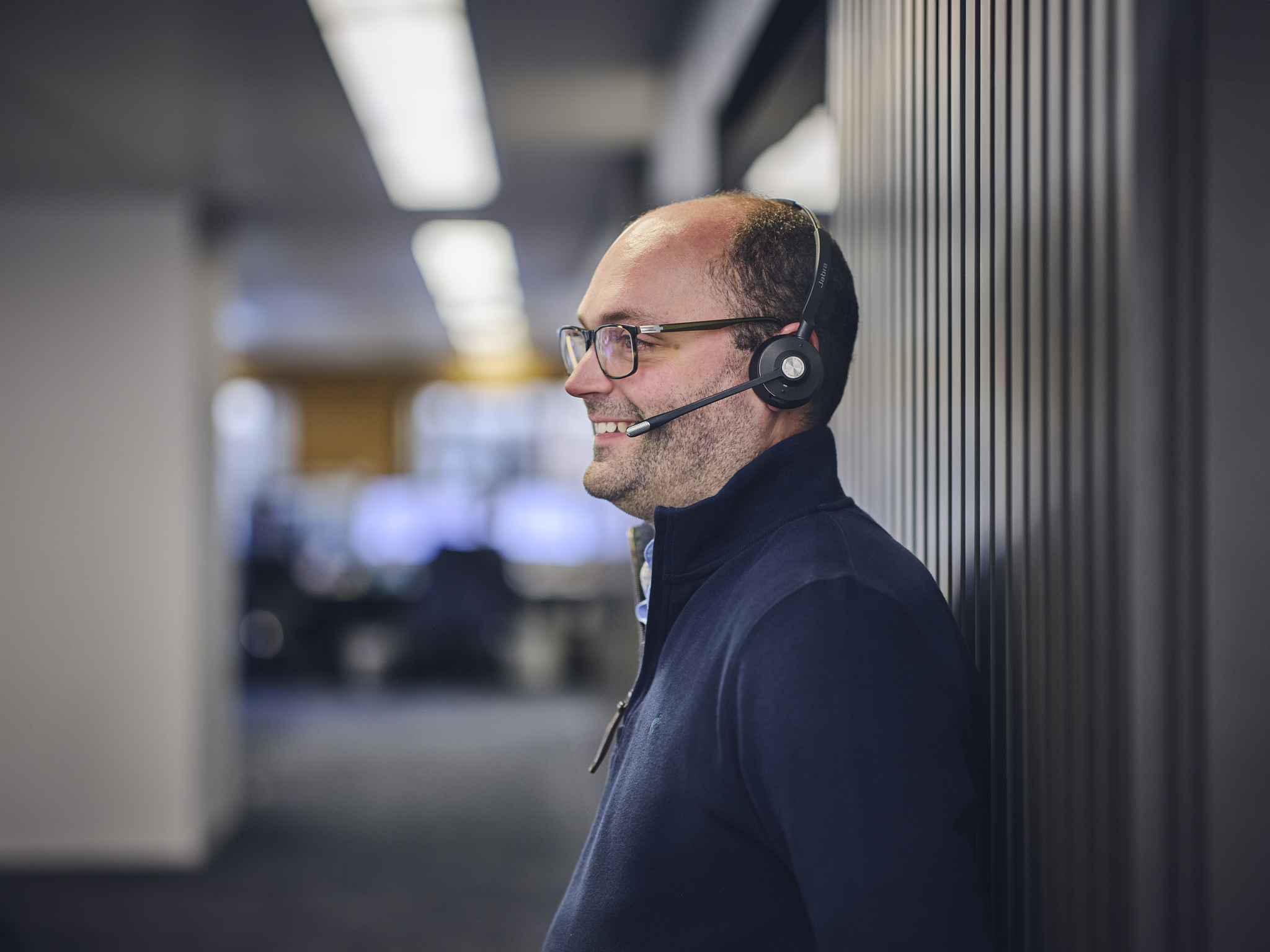Smiling man with glasses and a headset in an office environment.