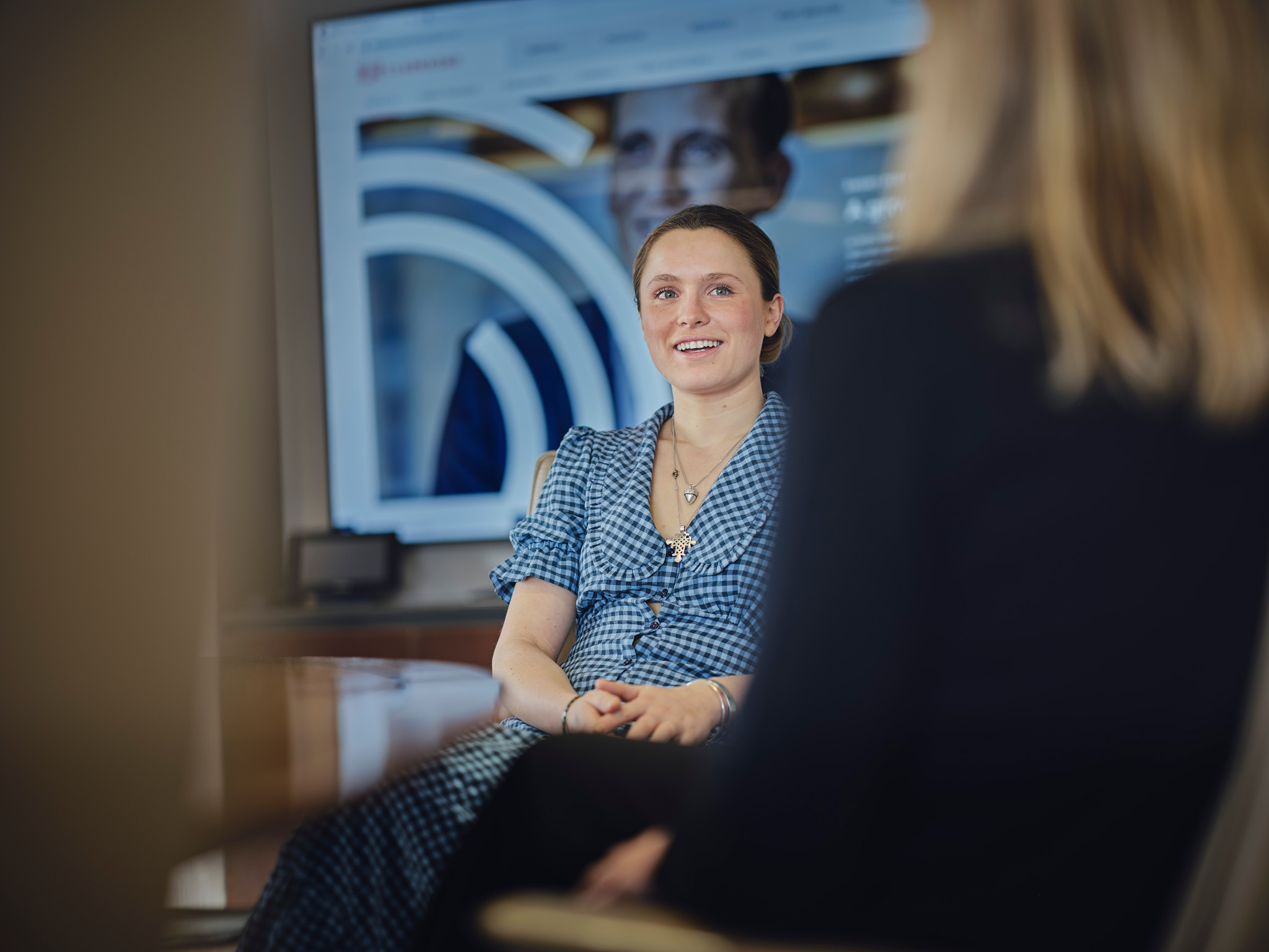 Smiling young woman with light brown hair in a checkered blouse seated at a table.