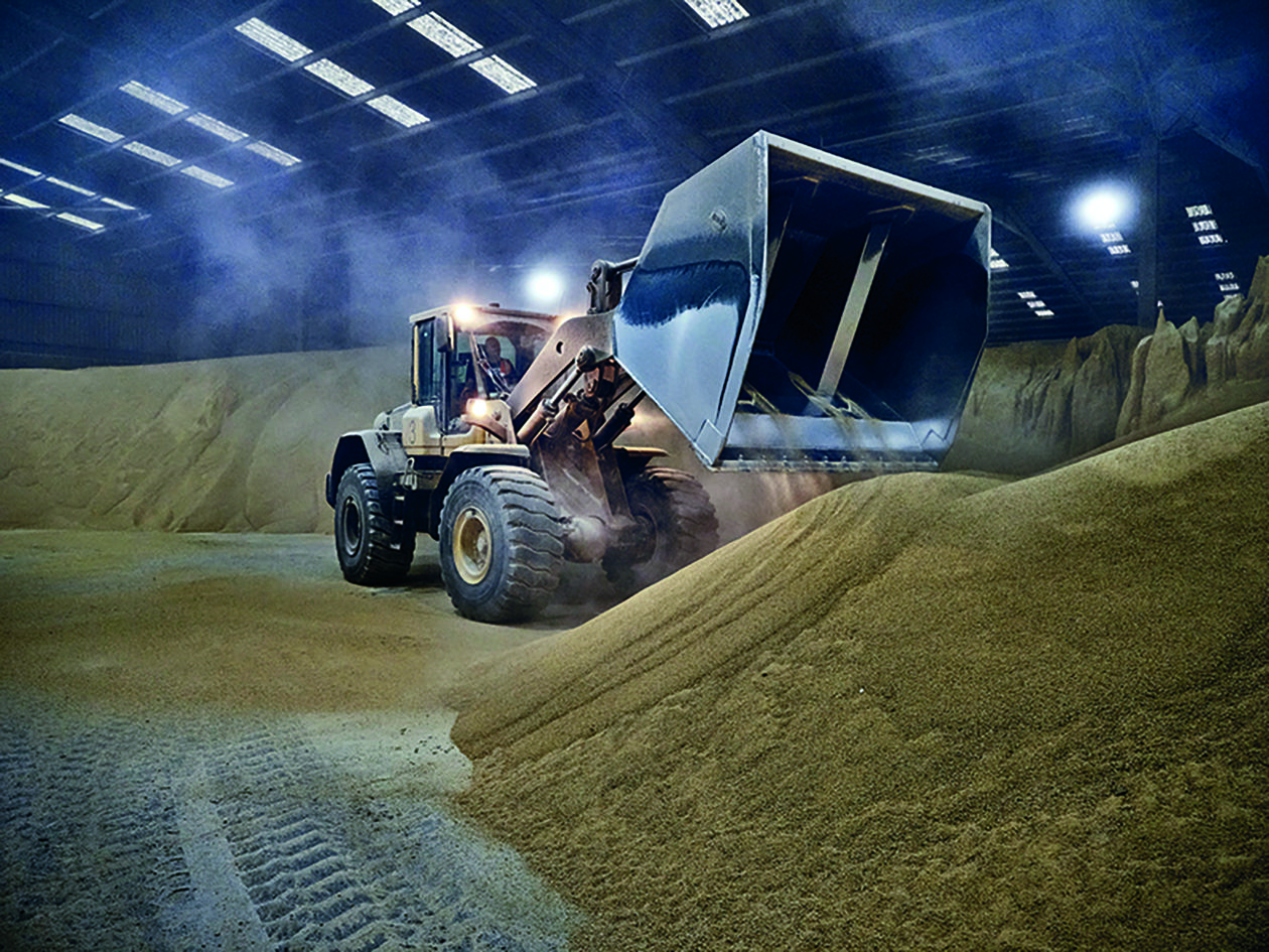 Large wheel loader operating inside a dimly lit warehouse, moving a pile of grain.