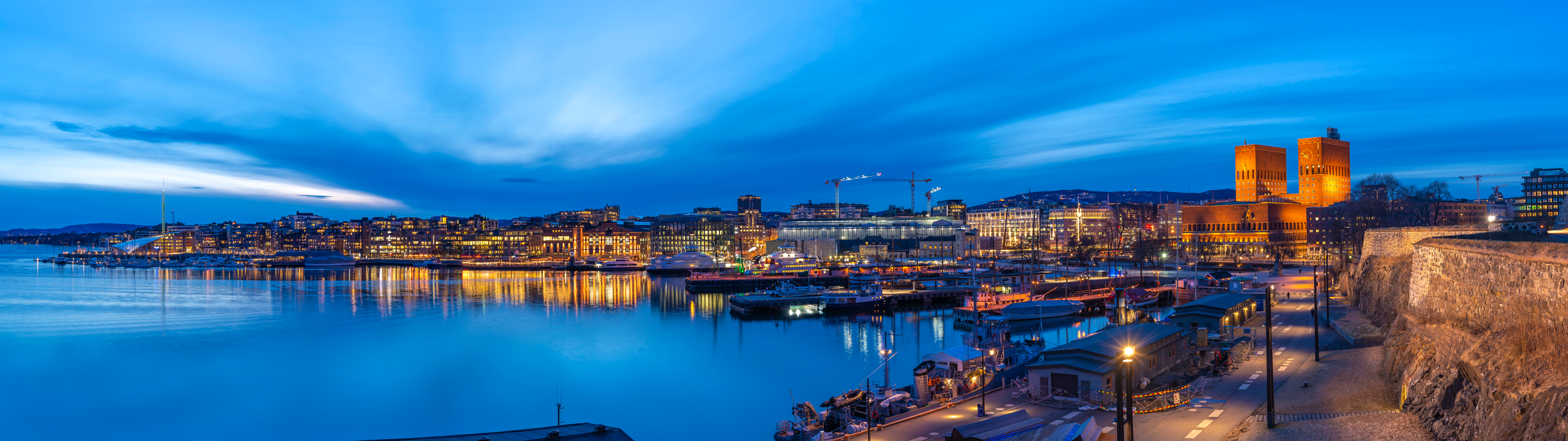 Panoramic view of a harbor city at dusk with modern waterfront buildings, reflections, and boats.
