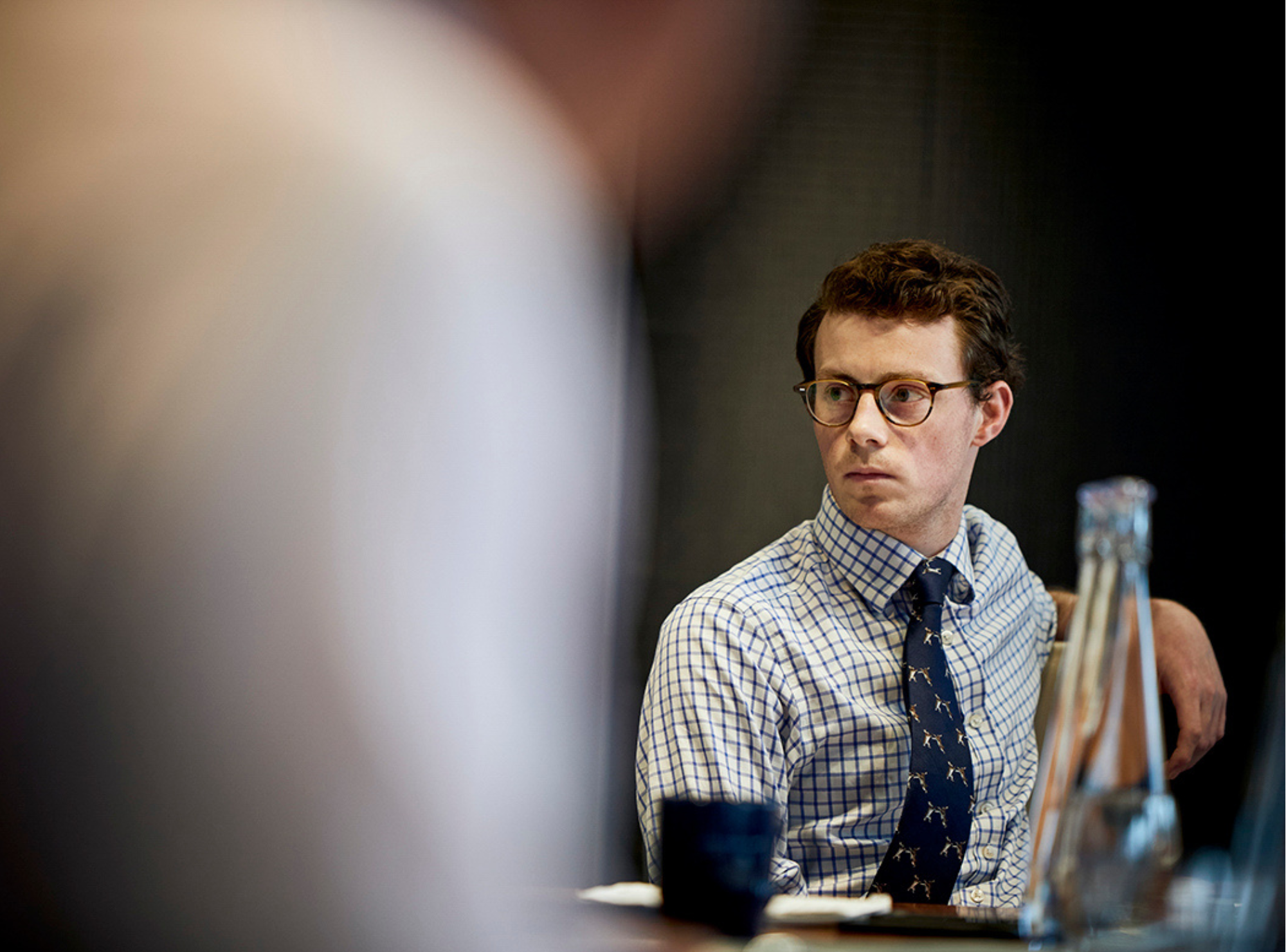 Man in checked shirt and tie, seated, looking serious.