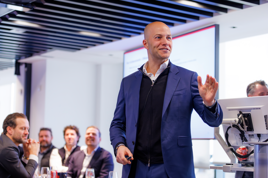 Smiling man in a blue blazer speaking and gesturing to a seated audience.
