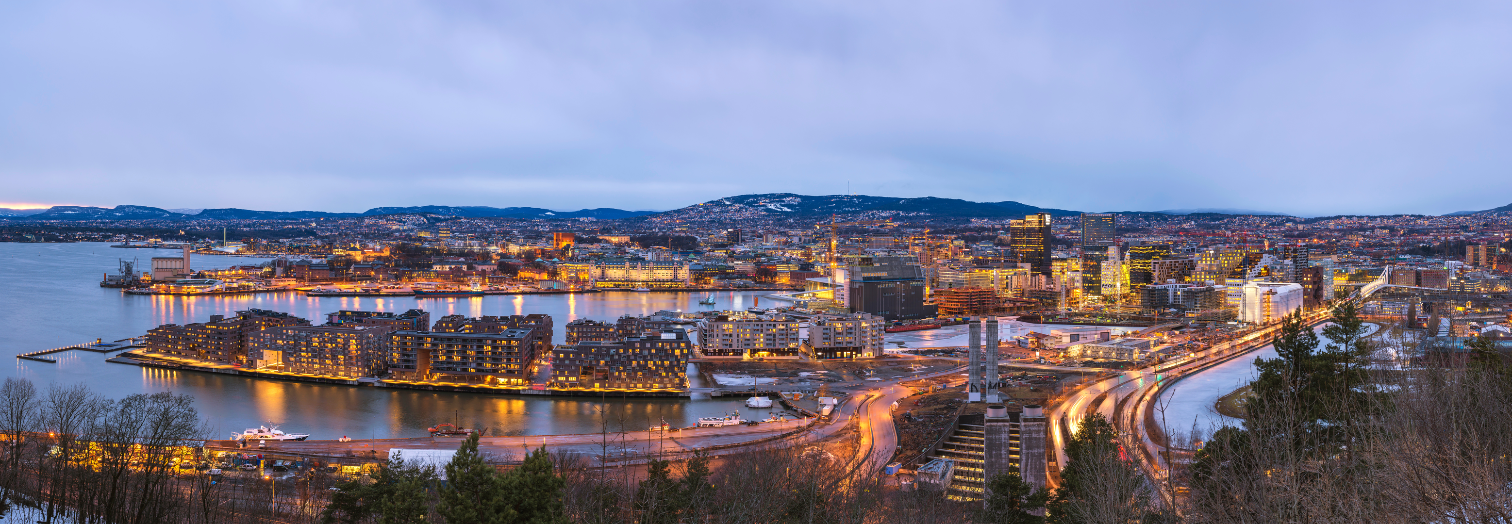 Panoramic twilight view of a city with illuminated buildings, water, and boats.