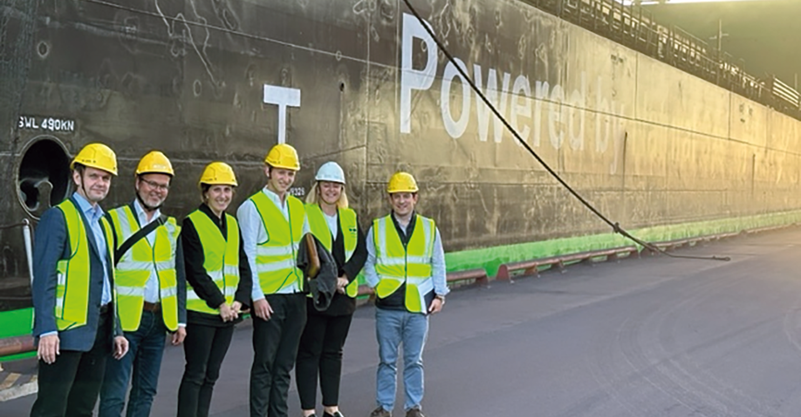 Group of six in hard hats and vests standing on a dock in front of a large ship.