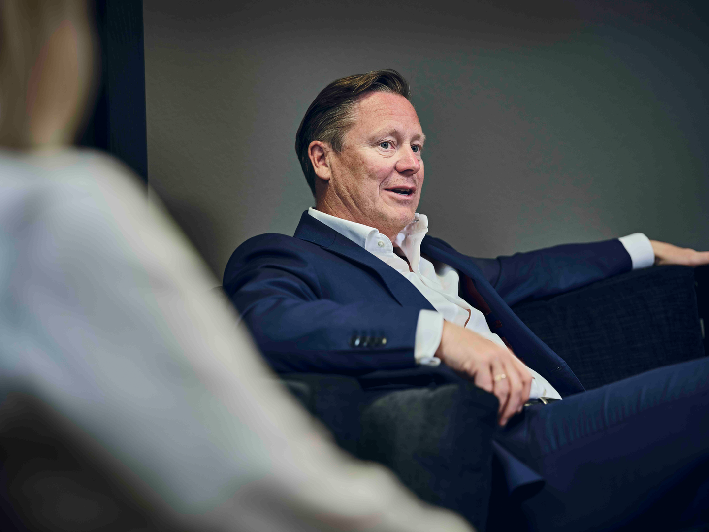 Man in a dark suit seated on a couch, engaging in conversation.