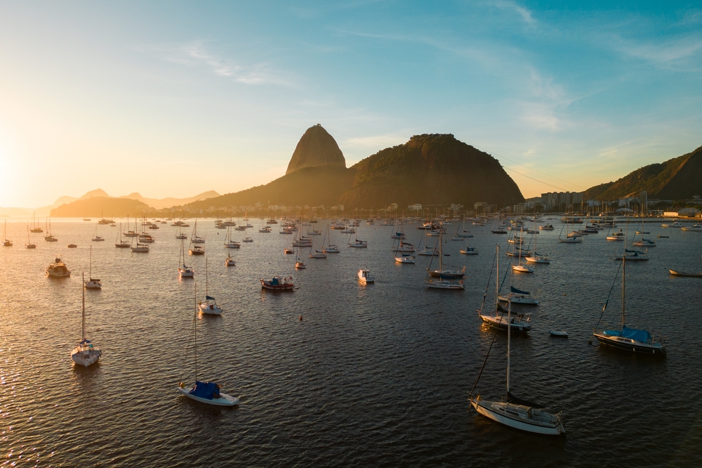 Serene sea landscape at sunset with boats, calm water, and mountains against a colourful sky.