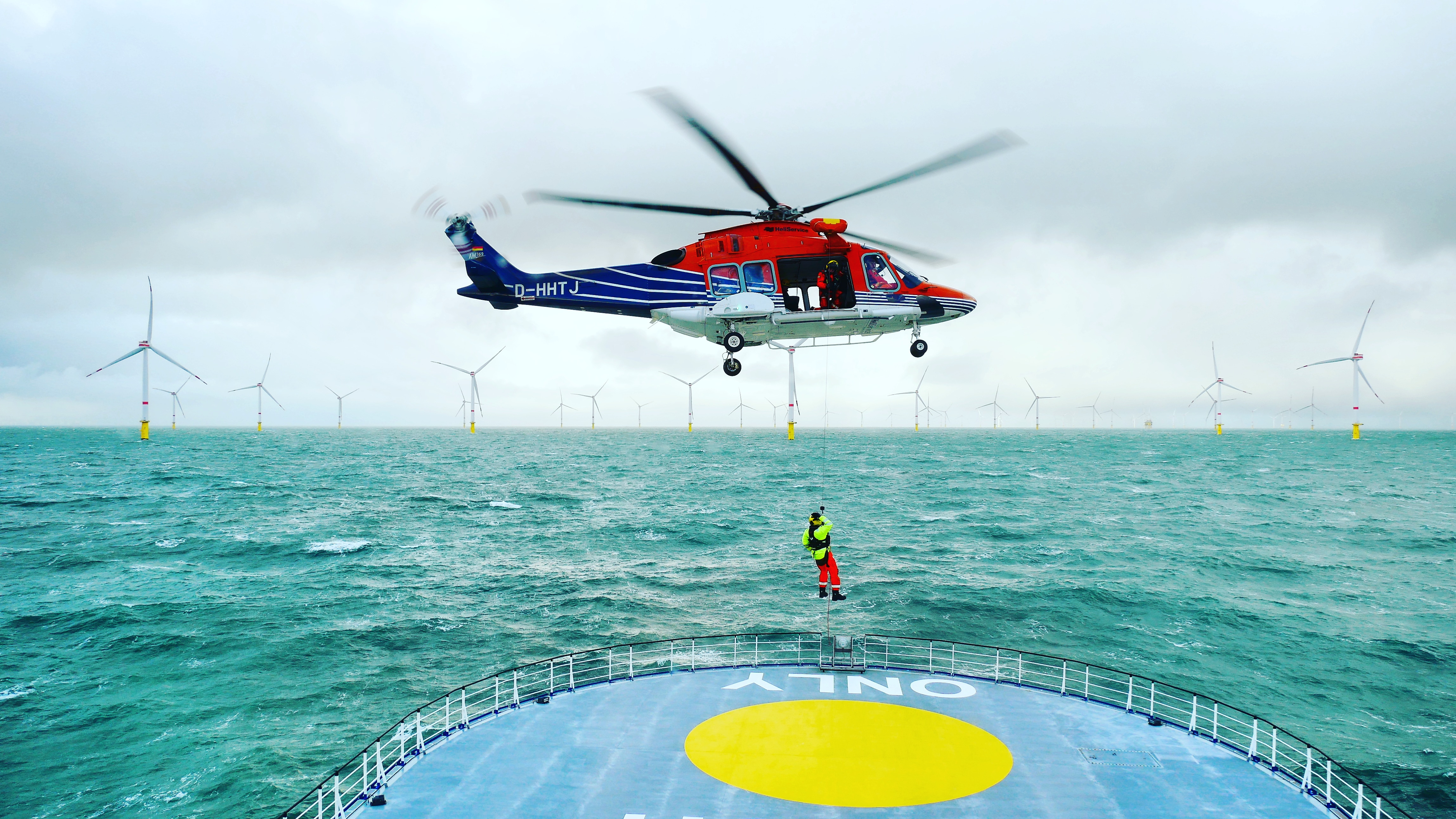 Helicopter hovering above a ship near wind turbines.